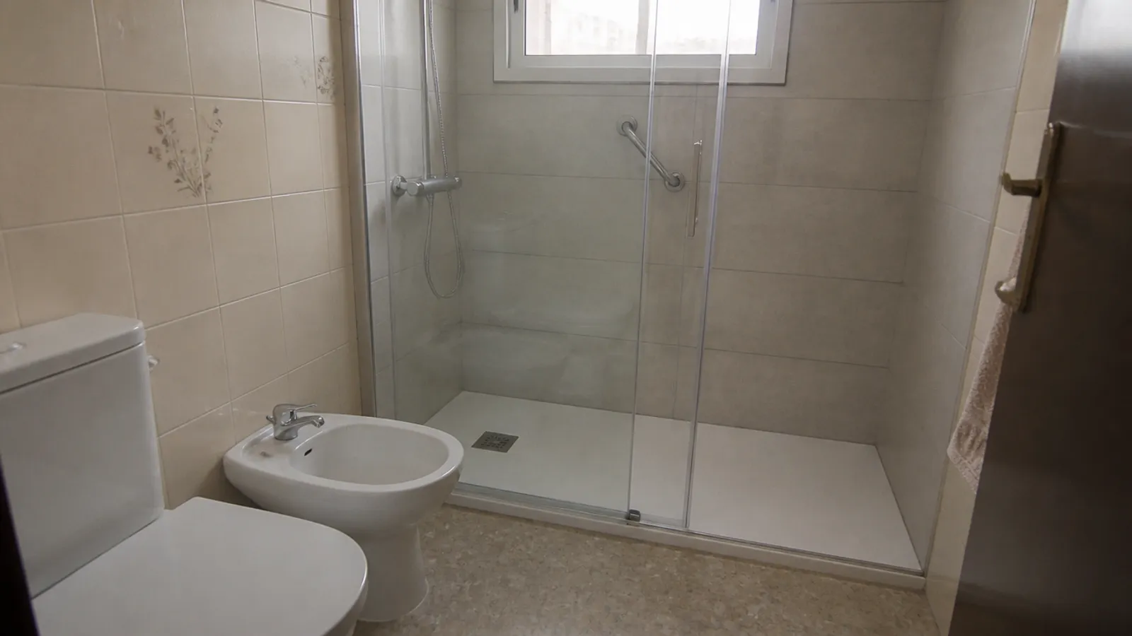 Plato de ducha extraplano blanco con mampara y asidero en un baño reformado de piso antiguo